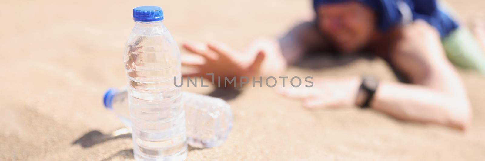 Close-up of man lying on sand in desert and feeling thirst. Guy reaching for bottle of water in sands. Dehydration under baking sun and water conservation idea