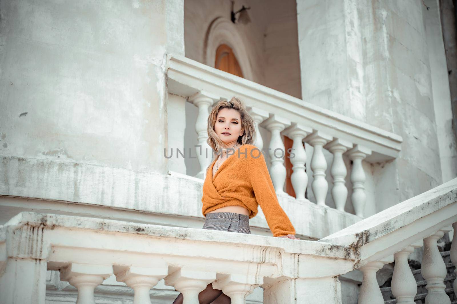 A middle-aged woman looks like a good blonde with curly beautiful hair and makeup on the background of the building. She is wearing a yellow sweater. by Matiunina