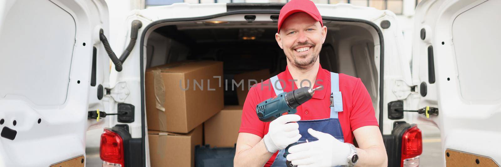 Smiling worker hold screwdriver device in hand in front of truck full of boxes by kuprevich