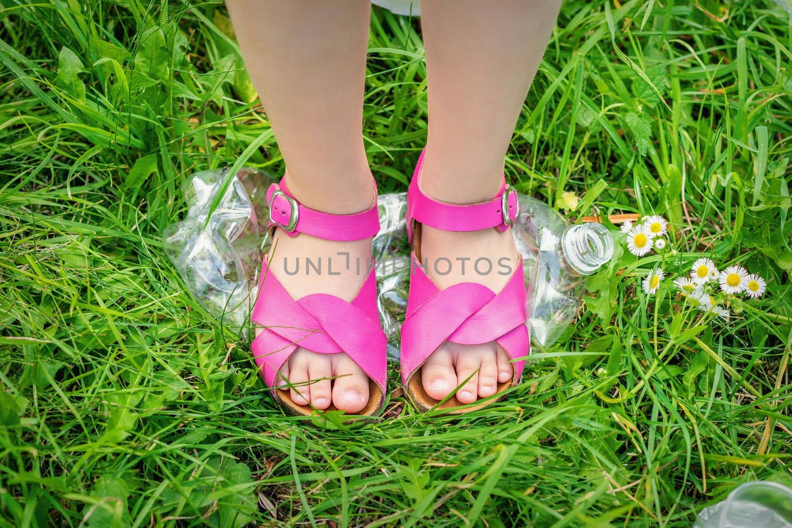 Legs of a little girl tramples a plastic bottle on the green grass in the park