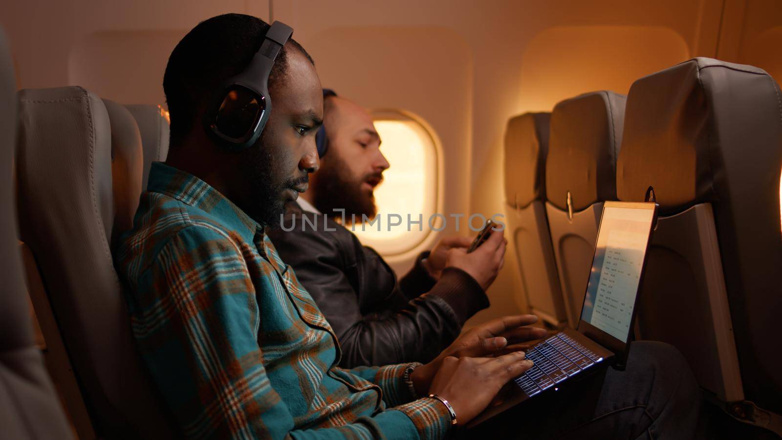 African american freelancer using laptop on flight, travelling on work trip during sunset in economy class. Working on computer and flying on airplane, aviation journey.