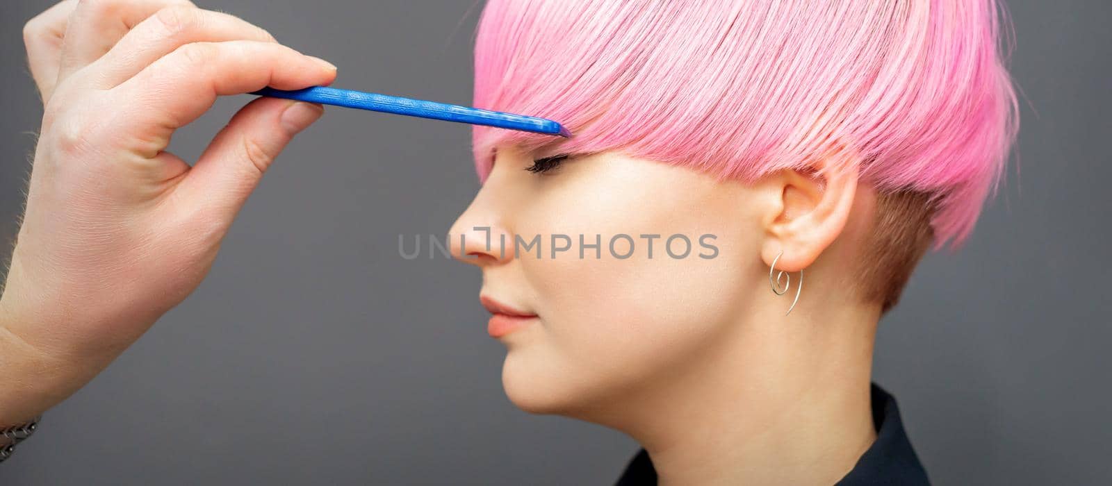 Hairdresser checking short pink hairstyle of young woman with comb on gray background