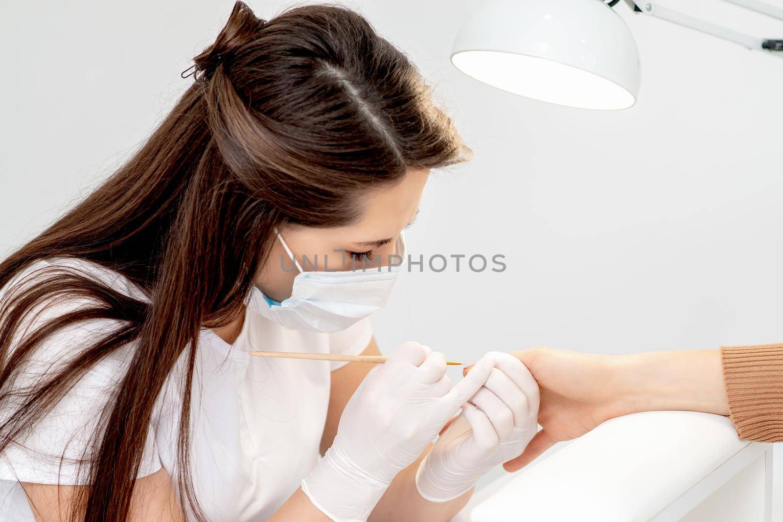 Manicure master with protective mask in rubber gloves applying beige nail polish on female nails in beauty salon