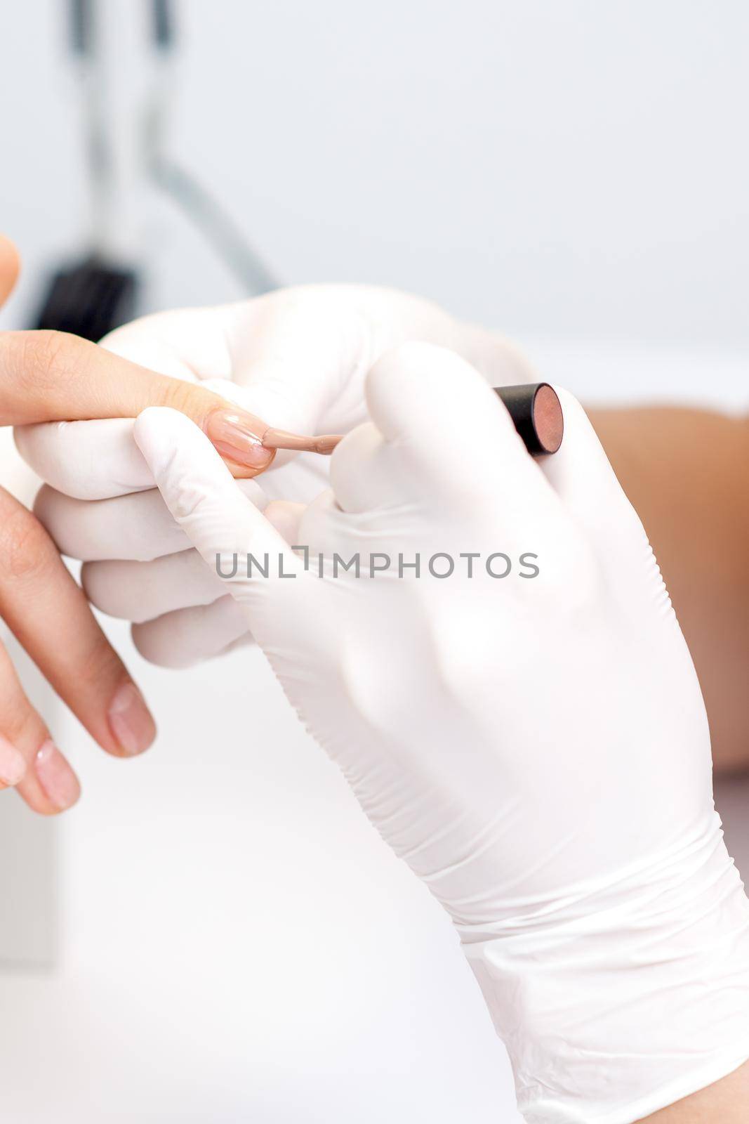 Manicure master in protective gloves applying beige nail polish on female nails in beauty salon