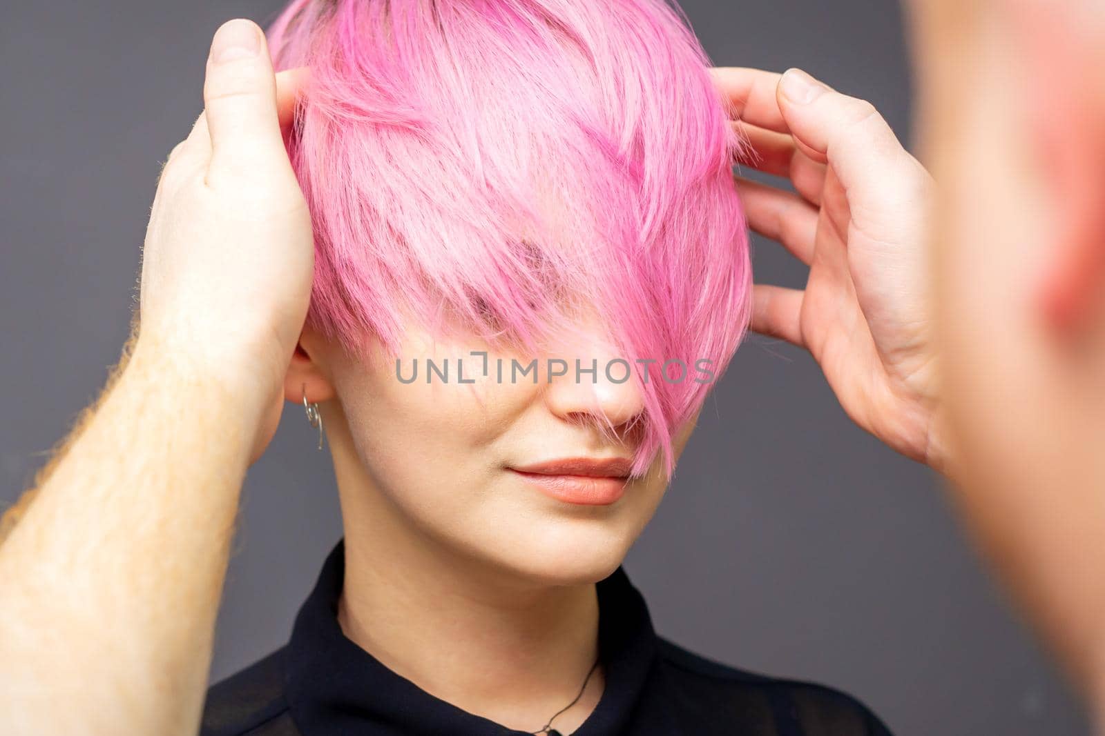 Hairdresser checking short pink hairstyle of young woman on gray background