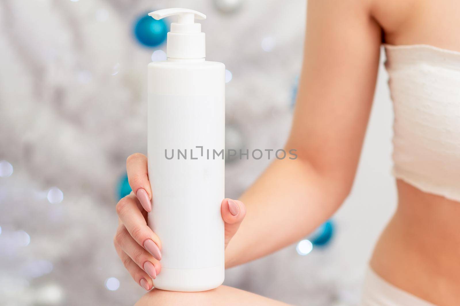 Cosmetics white bottle in hand of young female on christmas tree background. Concept of New Year discounts
