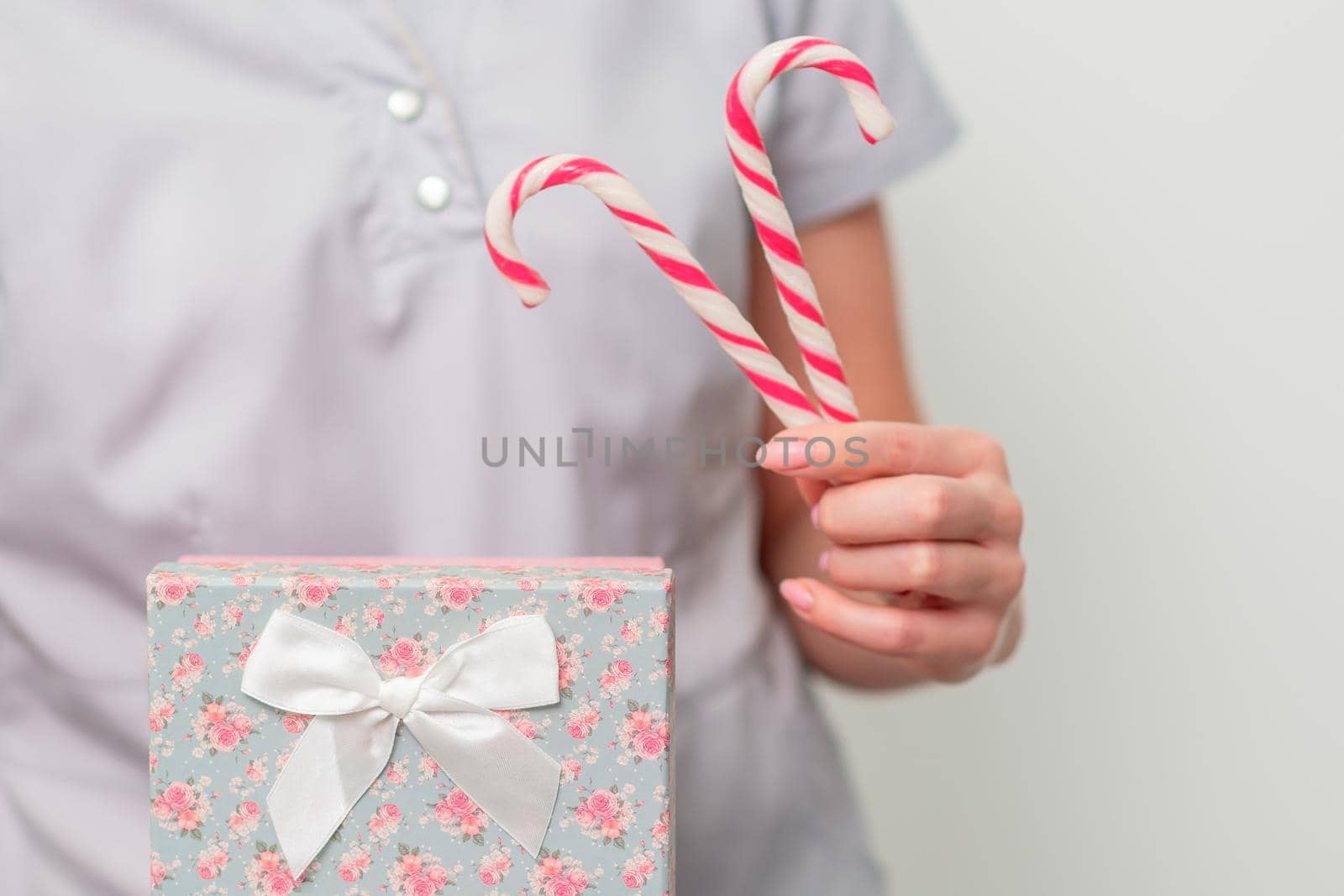 Young female in medical uniform with gift box and christmas candy near standing on light background