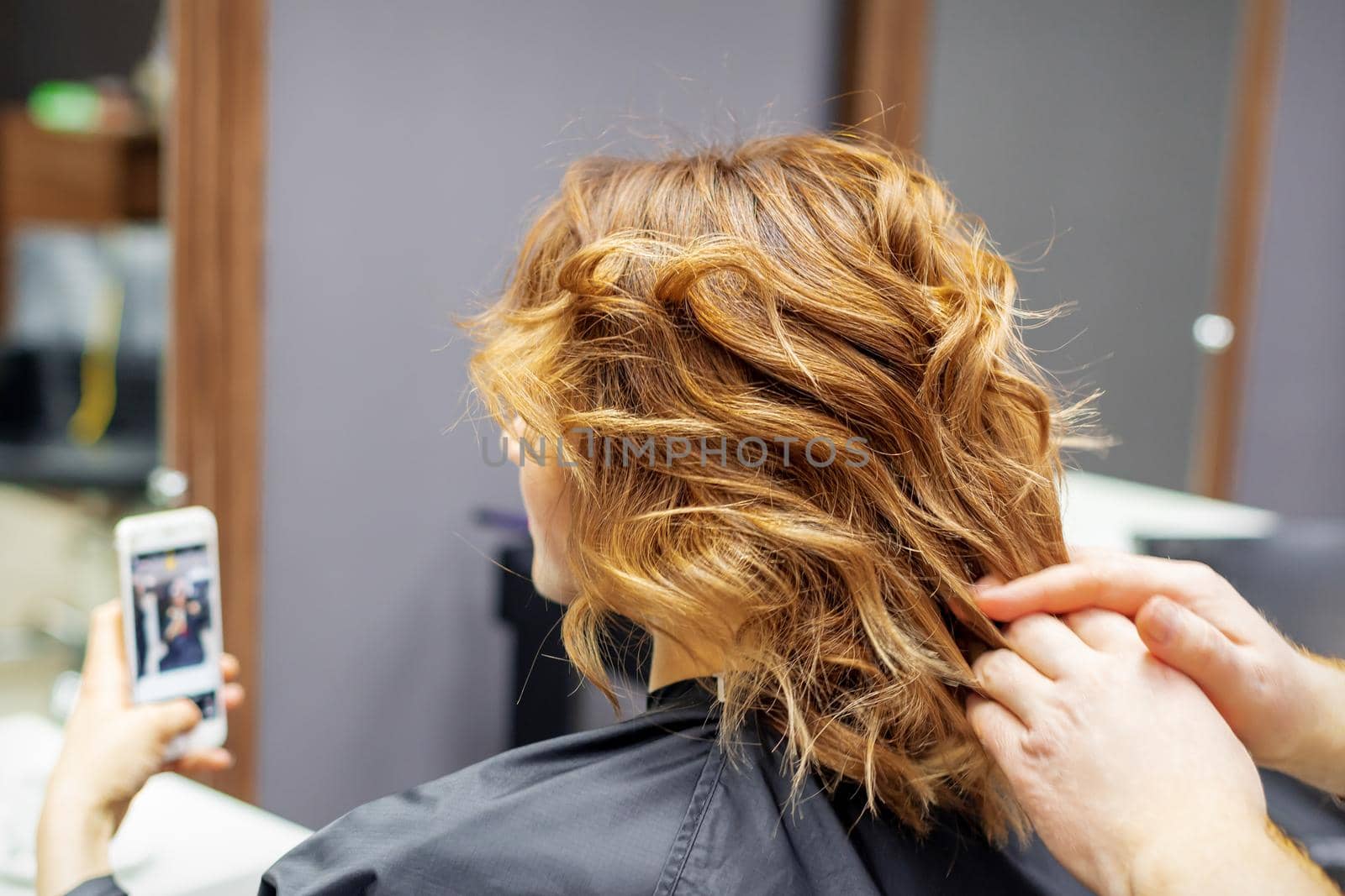 Hairdresser doing hairstyle for young woman with red curly hair and with smartphone in her hands in beauty salon