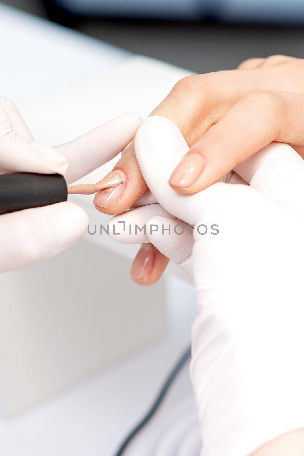 Manicure master in protective gloves applying beige nail polish on female nails in beauty salon