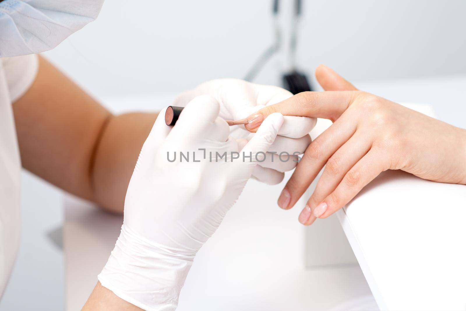 Manicure master in protective gloves applying beige nail polish on female nails in beauty salon