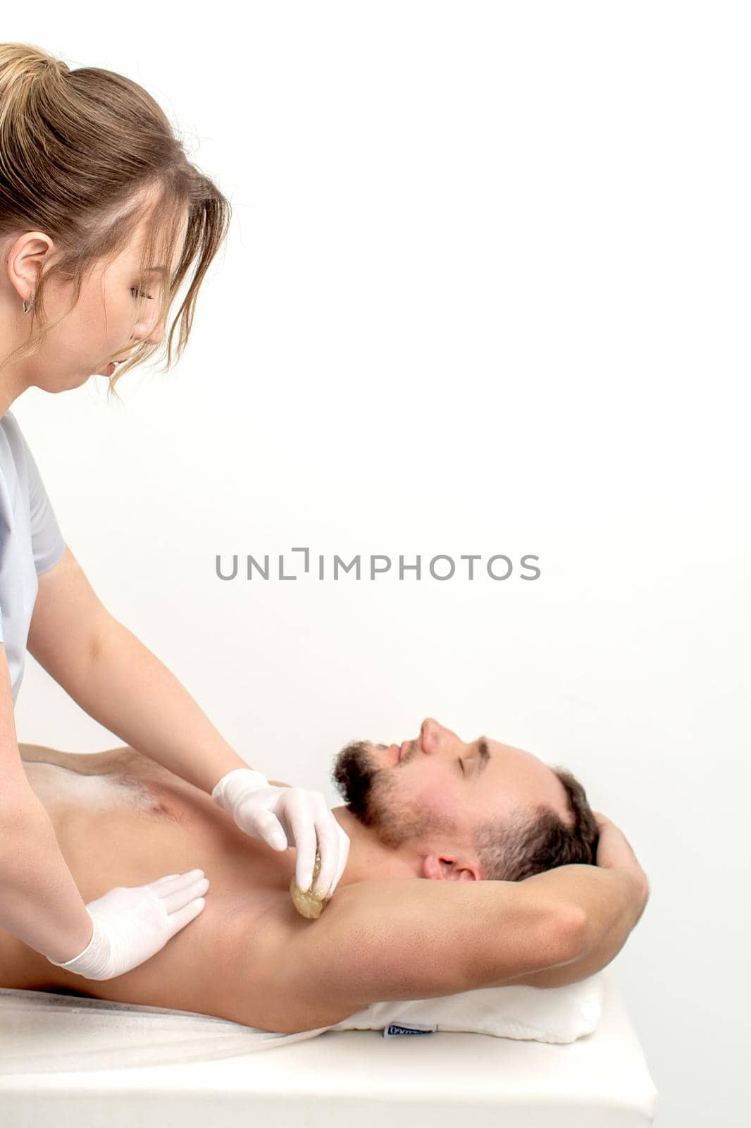 Young man receiving waxing underarm or epilation armpit by young female cosmetologist in beauty salon