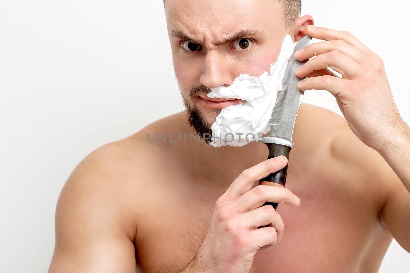Young caucasian man shaving beard with knife on white background