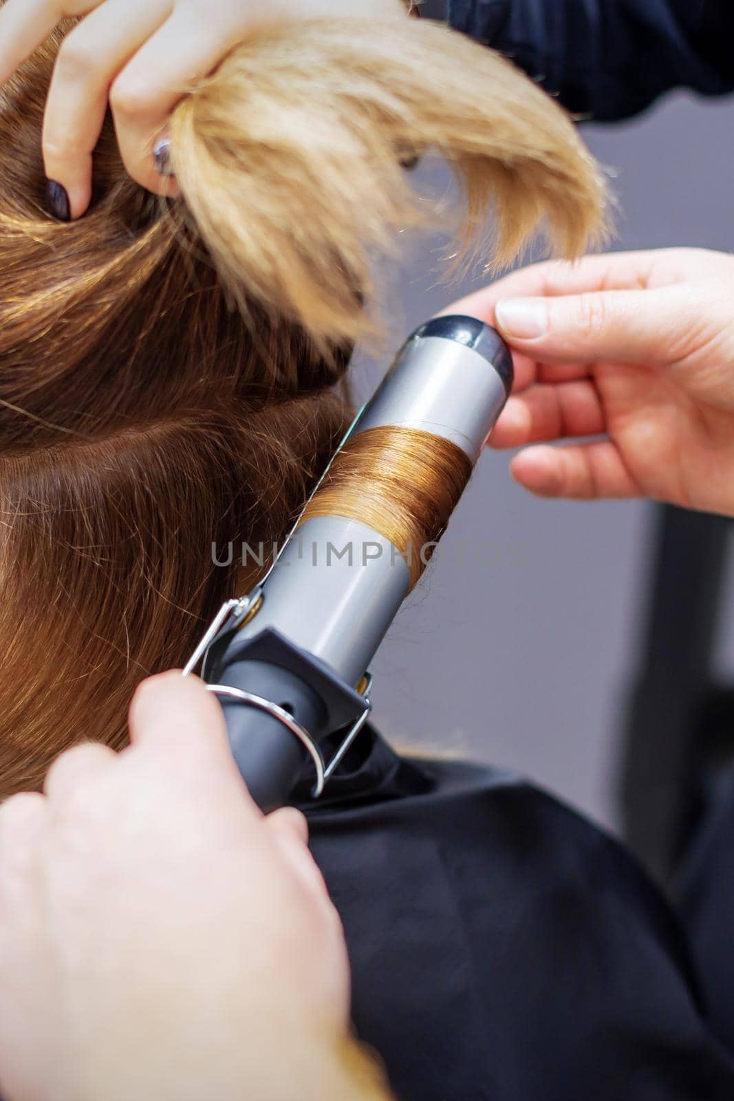 Professional hairdresser twists curls of long light brown hair of woman with curling iron in beauty salon. Hairdressing procedures