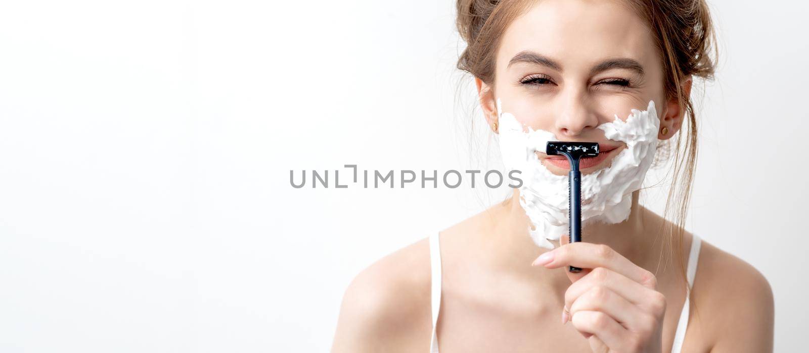 Beautiful young caucasian woman shaving her face by razor on white background. Pretty woman with shaving foam on her face