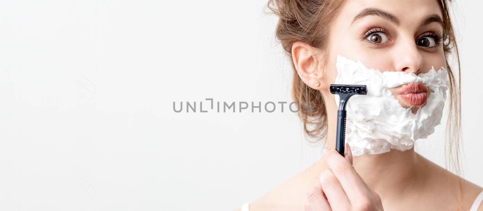 Beautiful young caucasian woman shaving her face by razor on white background. Pretty woman with shaving foam on her face