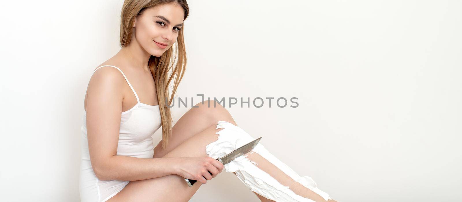 Beautiful young caucasian woman shaves her legs with a knife on a white background