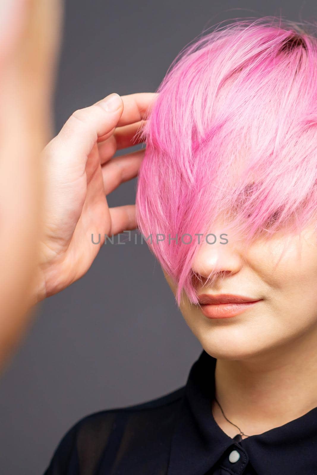 Hairdresser checking short pink hairstyle of young woman on gray background