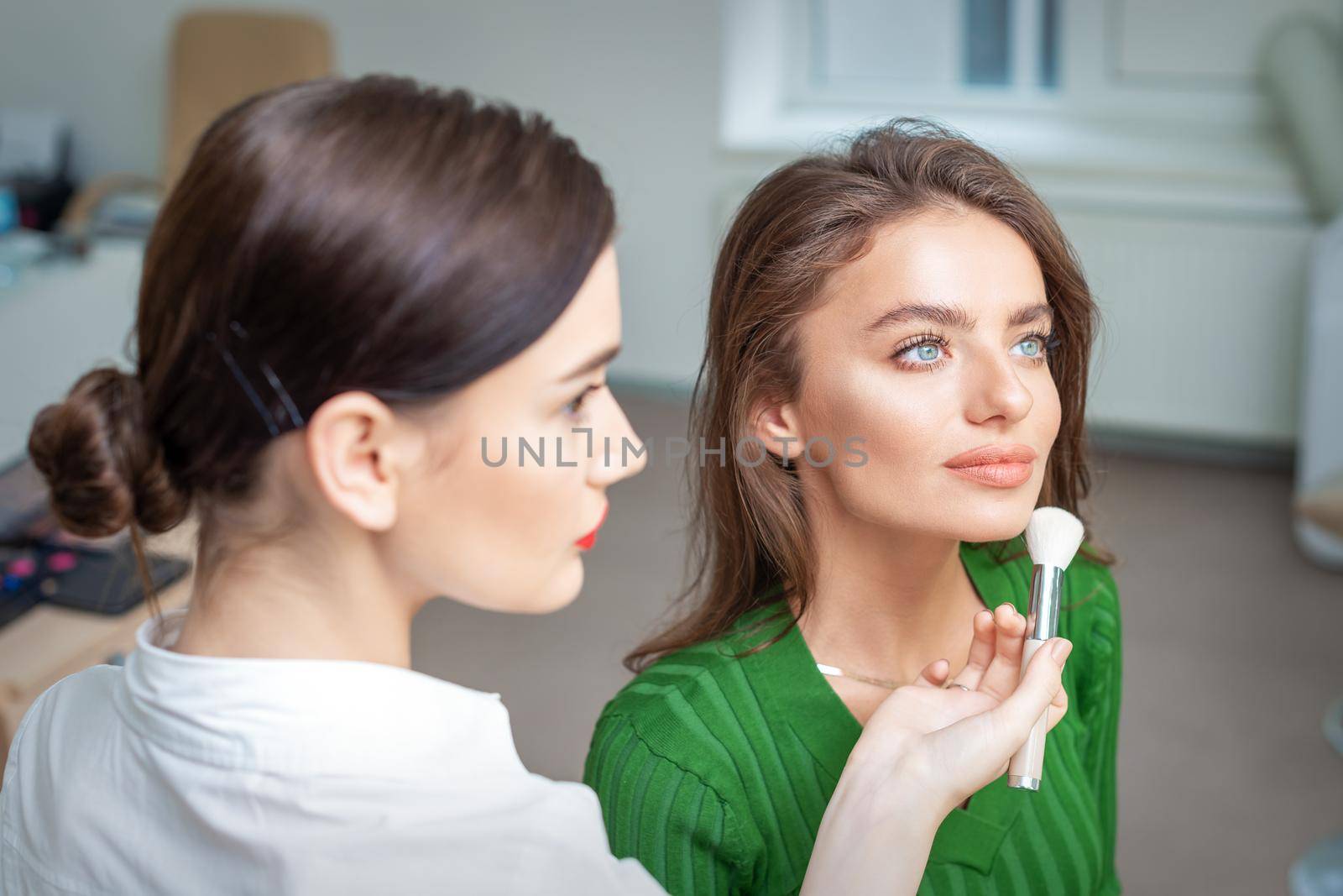 Make up artist applying professional make up of tonal foundation on the face of beautiful young caucasian woman in make up room. Base for make up.