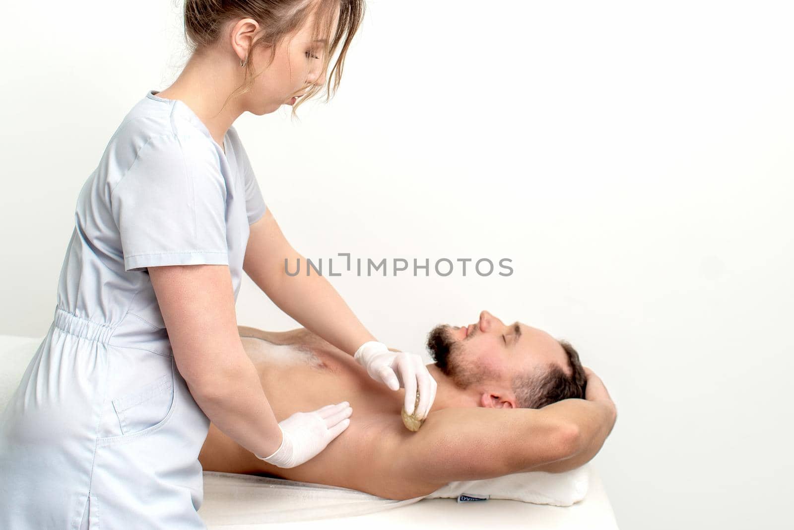 Young man receiving waxing underarm or epilation armpit by young female cosmetologist in beauty salon