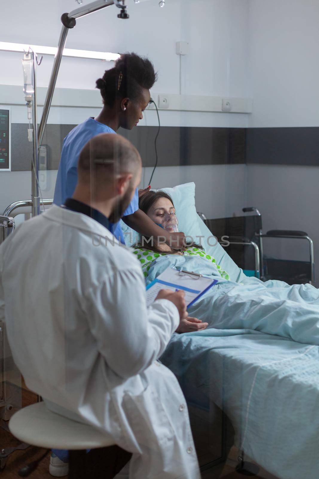 Practitioners team checking patient putting oxigen mask monitoring respiratory illness during coronavirus pandemic in hospital ward. Nurse with black skin checking heart puls using medical stethoscope