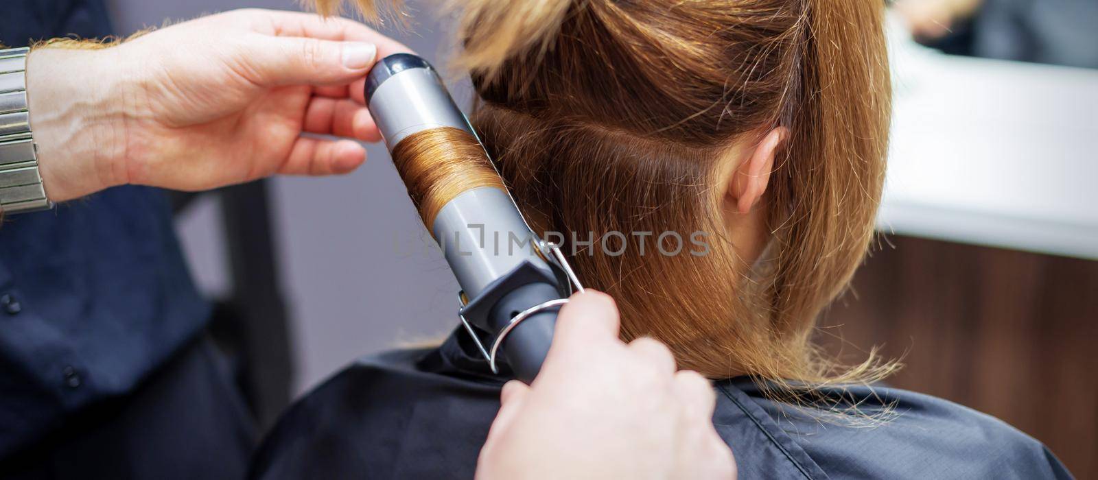 Professional hairdresser twists curls of long light brown hair of woman with curling iron in beauty salon. Hairdressing procedures