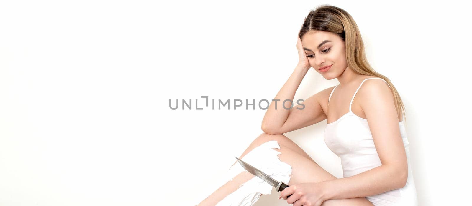 Beautiful young caucasian woman shaves her legs with a knife on a white background