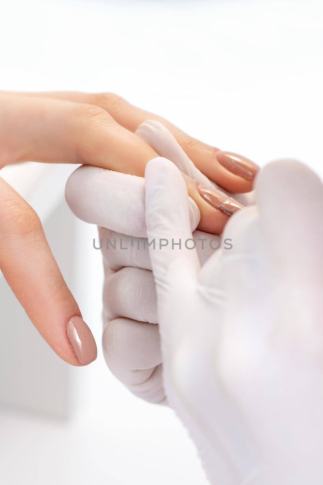Manicure master in protective gloves applying beige nail polish on female nails in beauty salon