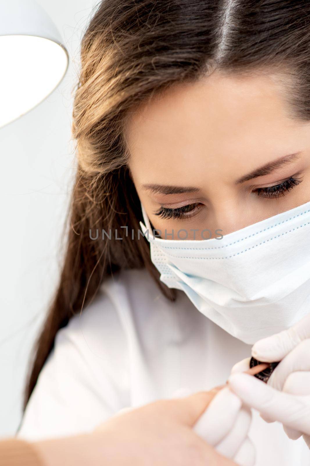 Manicure master with protective mask in rubber gloves applying beige nail polish on female nails in beauty salon