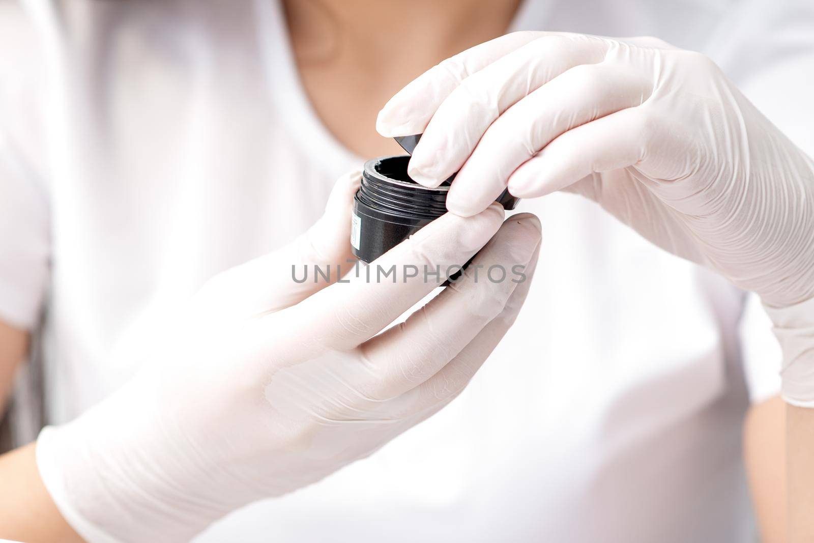 Beautician or manicure master with protective mask holds black jar with cosmetics or manicures product in her hands with white gloves