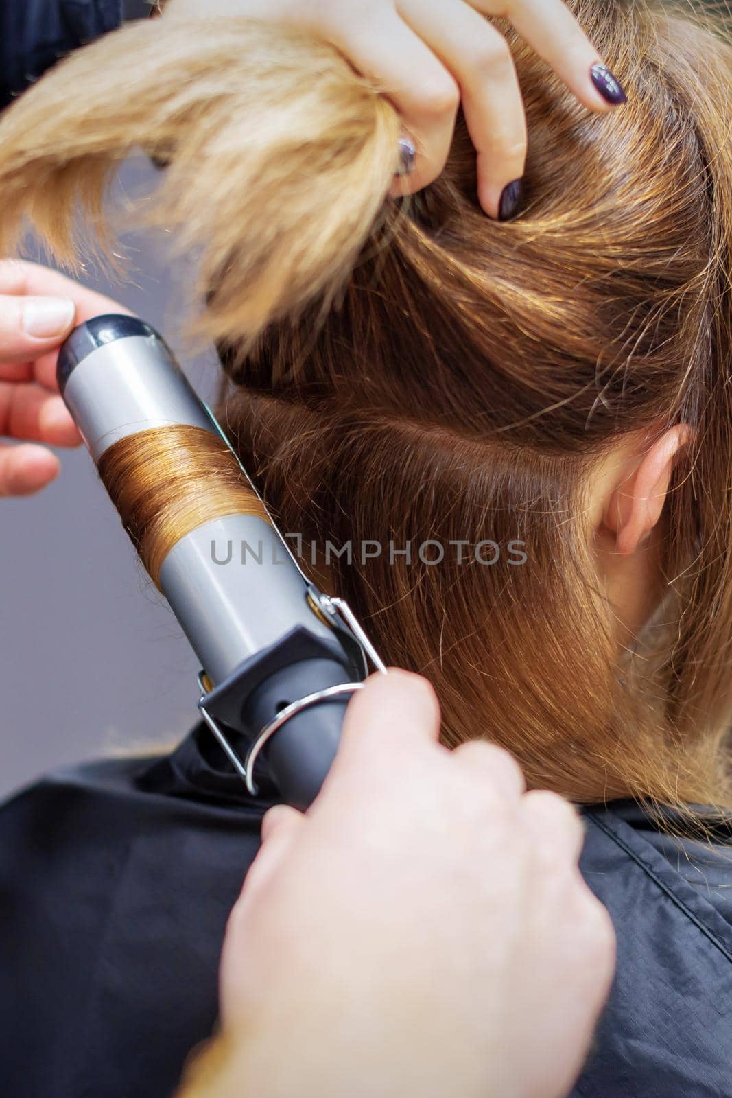 Professional hairdresser twists curls of long light brown hair of woman with curling iron in beauty salon. Hairdressing procedures