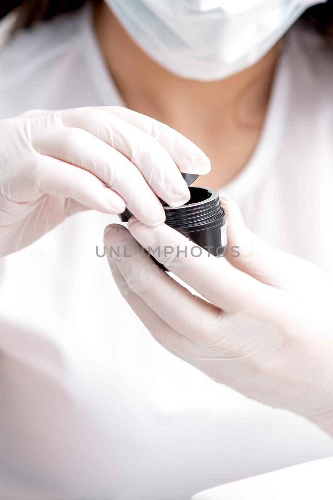 Beautician or manicure master with protective mask holds black jar with cosmetics or manicures product in her hands with white gloves