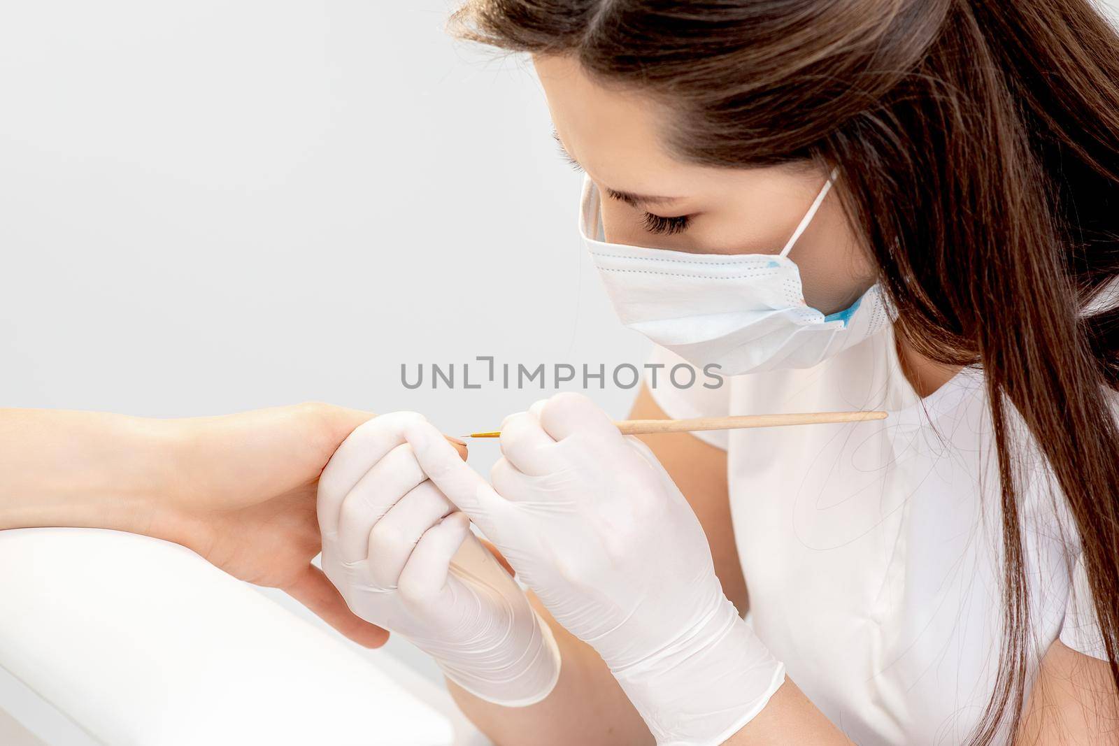 Manicure master with protective mask in rubber gloves applying beige nail polish on female nails in beauty salon