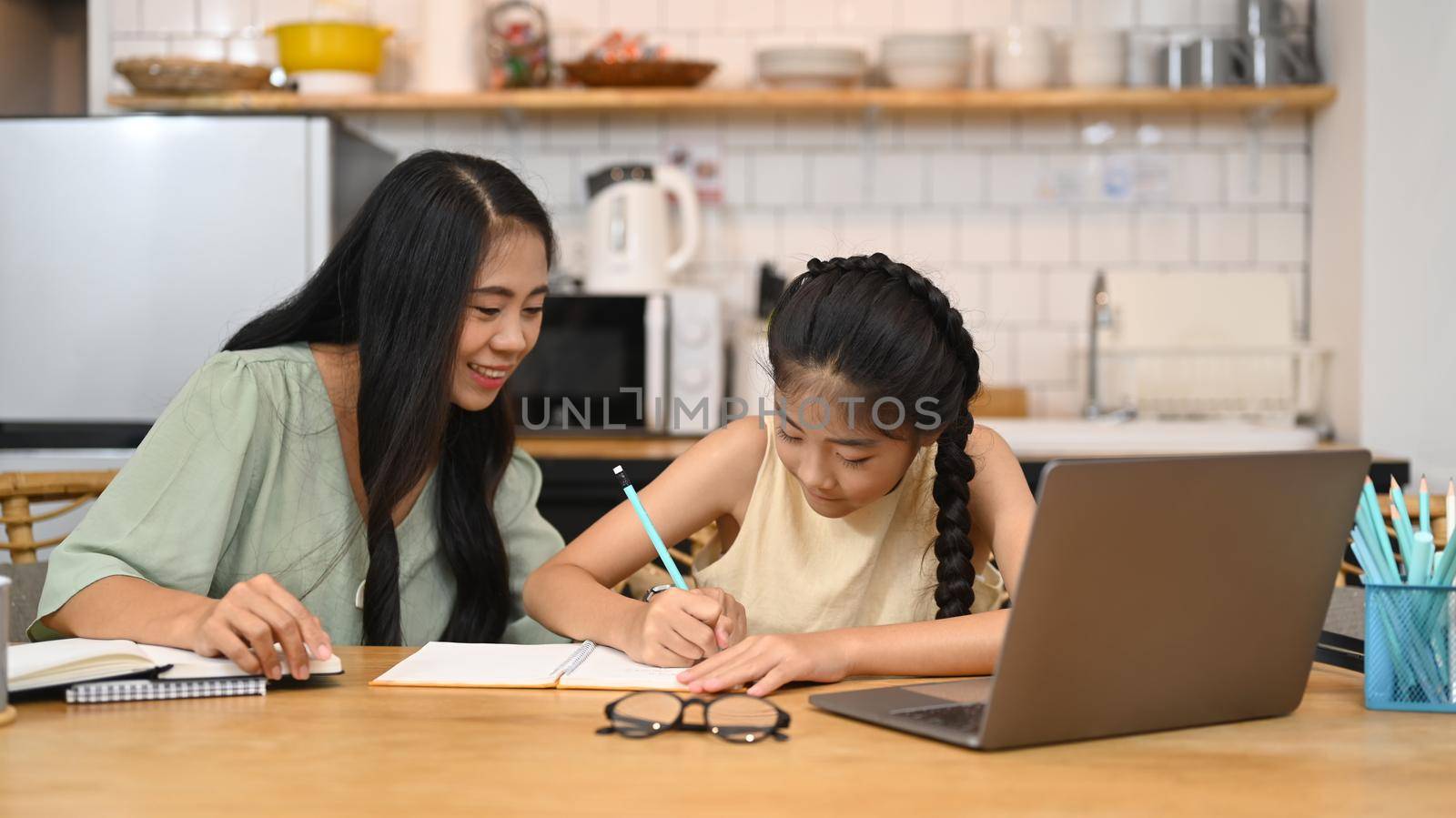 Asian mom helping her sweet daughter doing homework, learning online at virtual class on laptop computer at home.