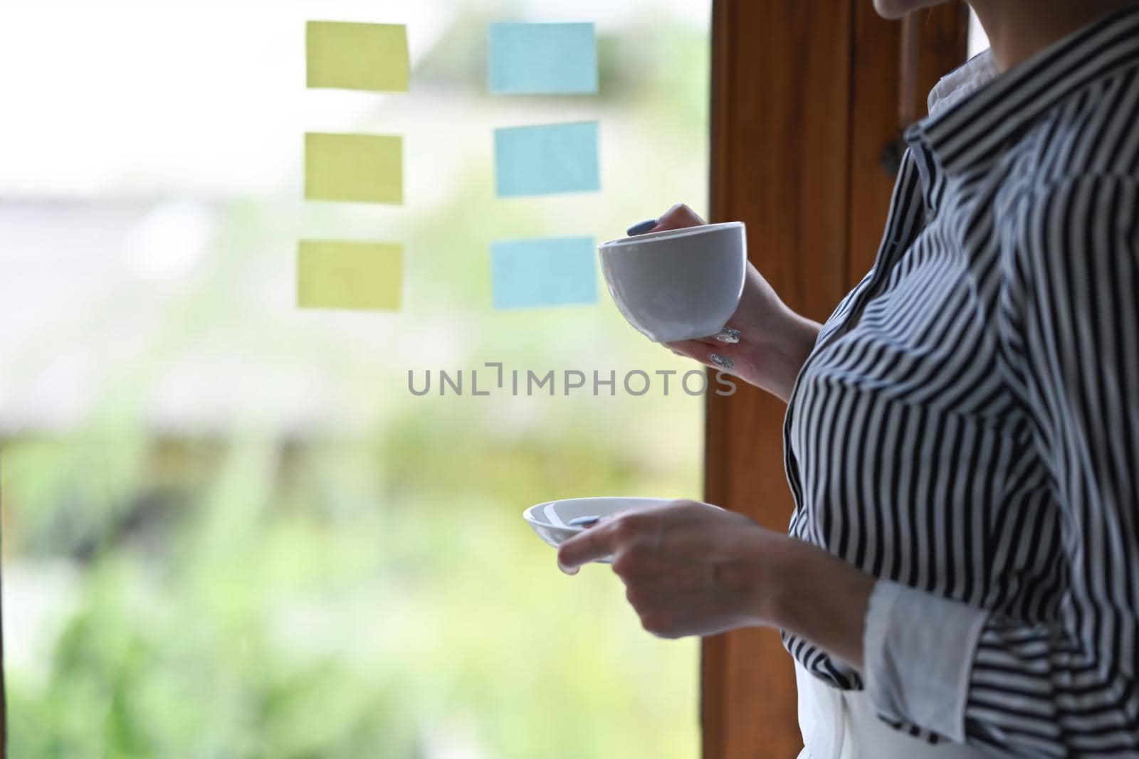 Businesswoman holding coffee cup and reading information on sticky notes at bright office.