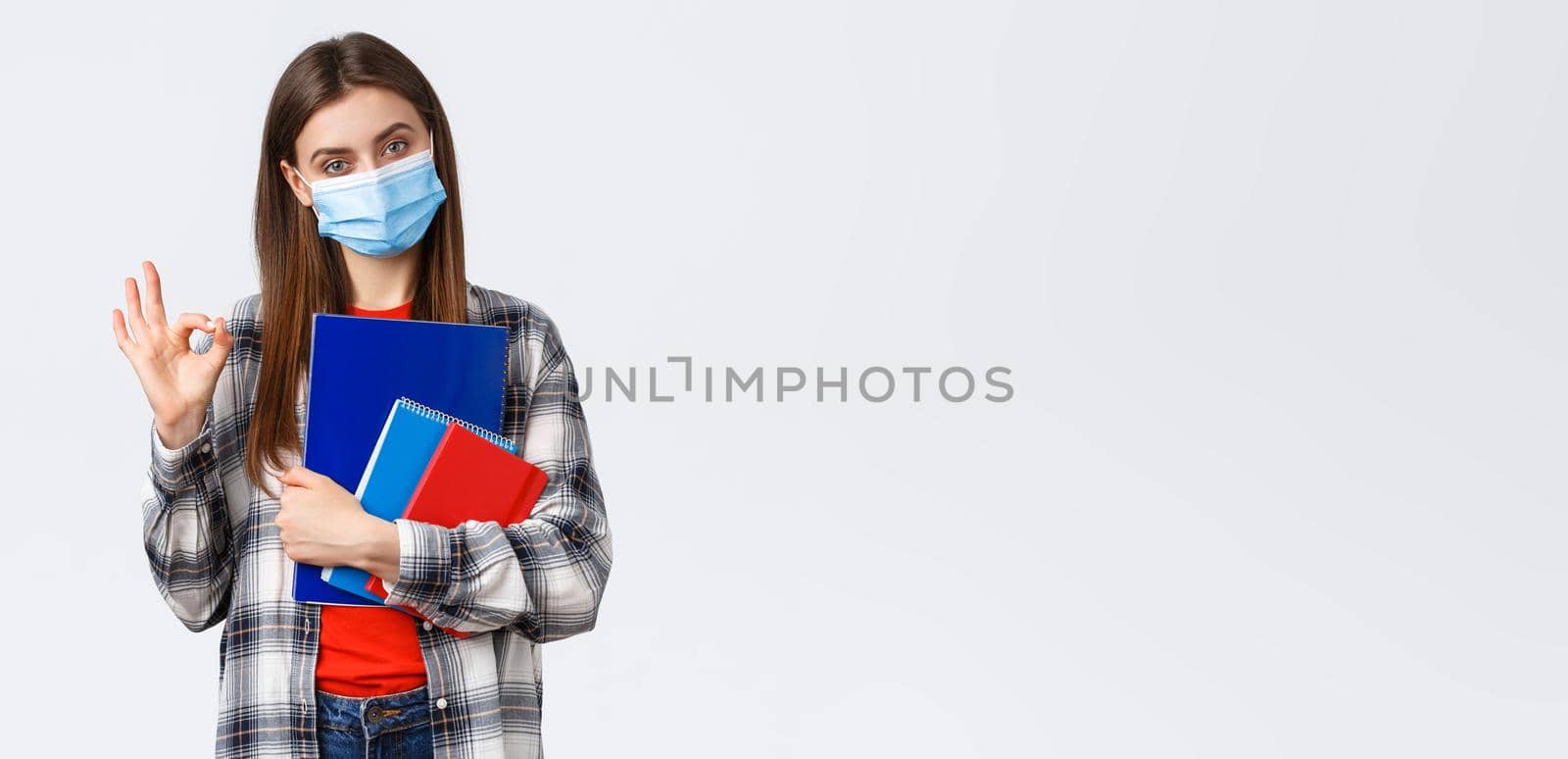 Coronavirus pandemic, covid-19 education, and back to school concept. Determined young woman, student in medical mask with notebooks and study material, show okay, no problem sign.