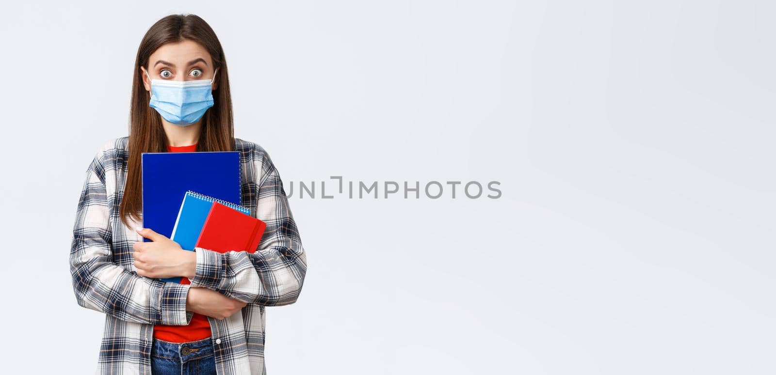 Coronavirus pandemic, covid-19 education, and back to school concept. Surprised female student in medical mask look amazed, widen eyes from big news, carry notebooks, white background.