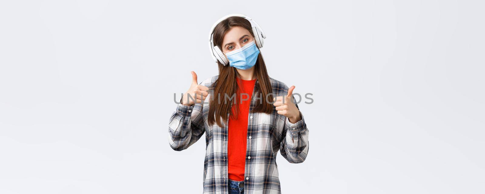 Social distancing, leisure and lifestyle on covid-19 outbreak, coronavirus concept. Pleased good-looking woman in medical mask, listening music headphones, show thumb-up good headsets.