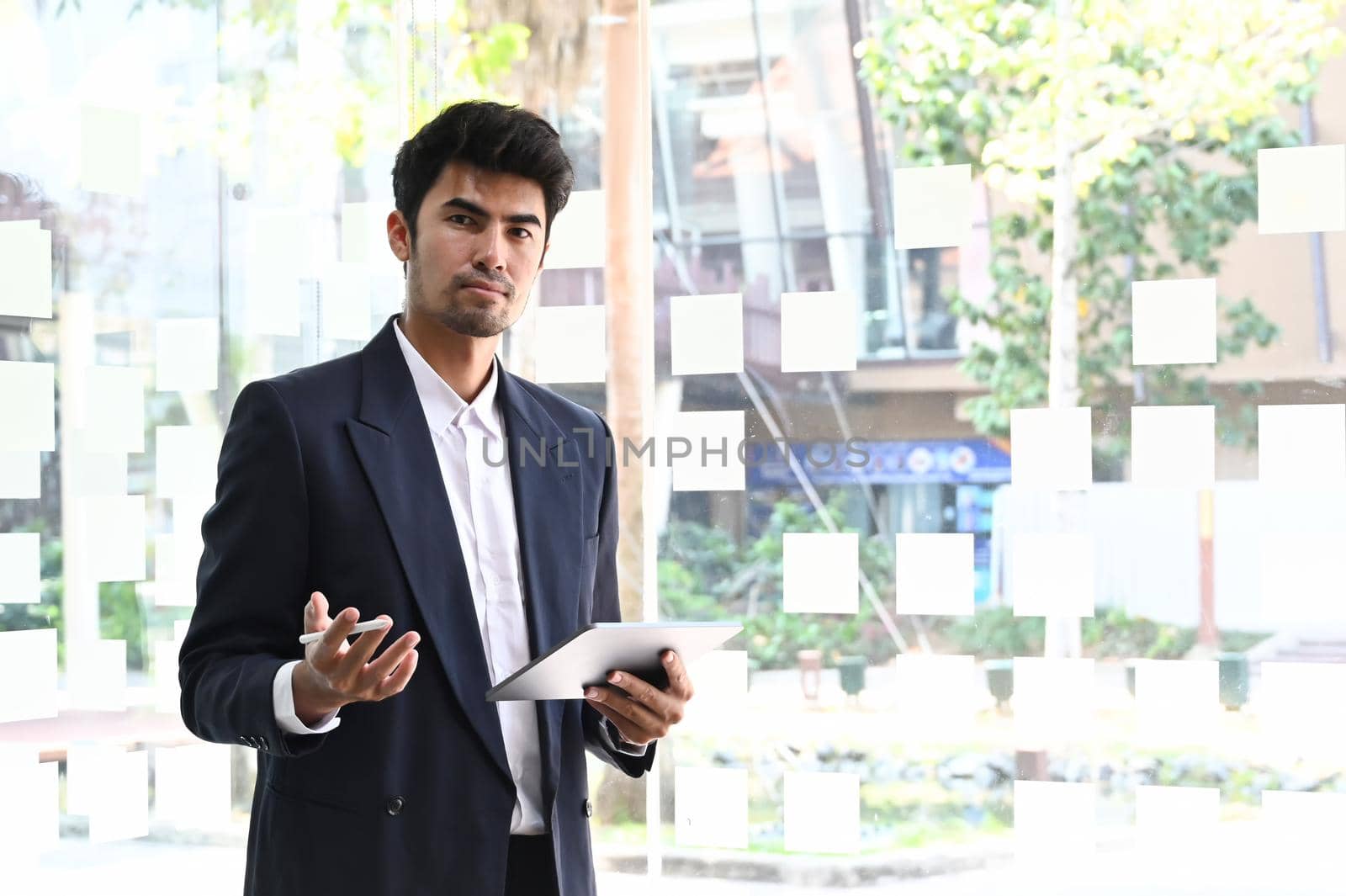 Confident professional businessman in black suit holding digital tablet and standing in bright modern office.