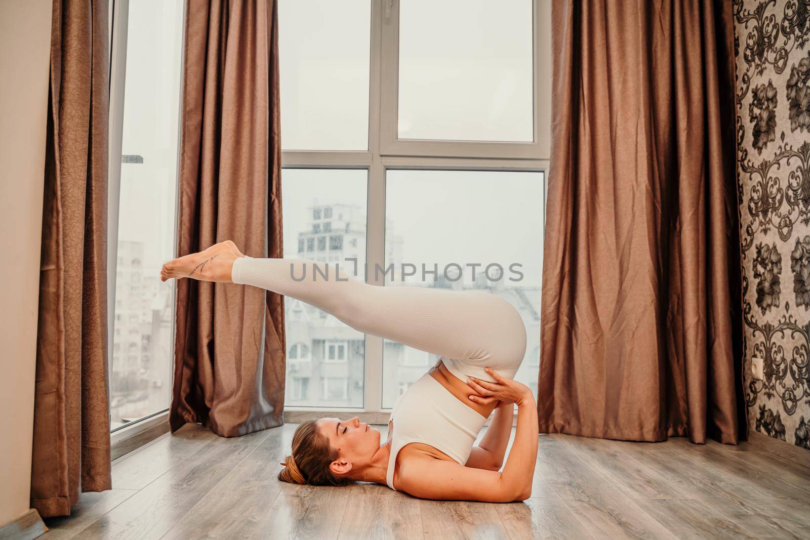 Young athletic attractive woman practicing yoga. Works out at home or in a yoga studio, sportswear, white pants and a full-length top indoors. Healthy lifestyle concept.