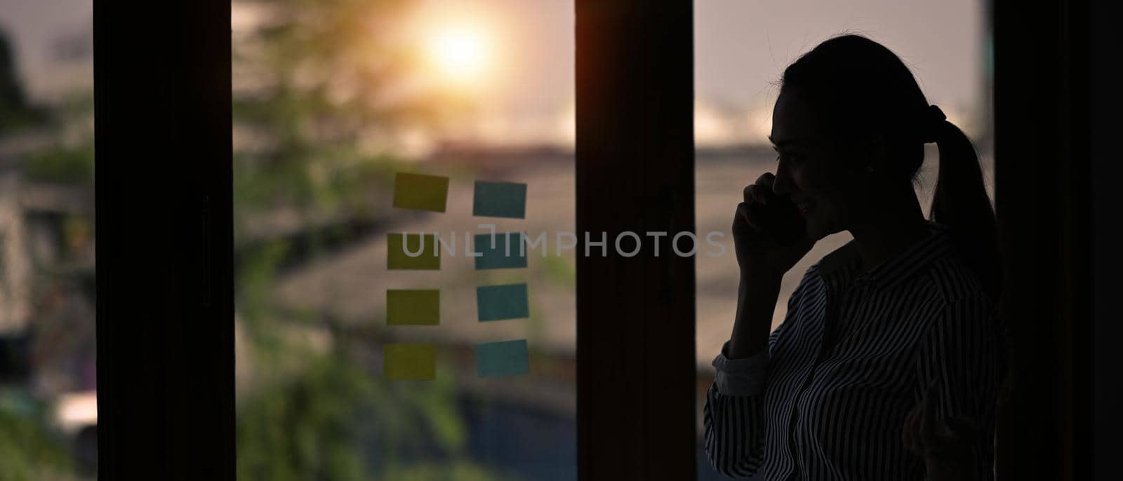 Silhouette of young woman talking on mobile phone while standing near window with view on sunset in city.