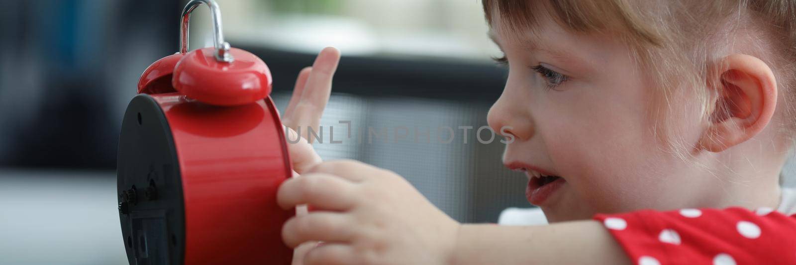 Portrait of curious little pretty girl explore red clock touch dial with finger. Kid interested in learning time and new skills. Education process concept