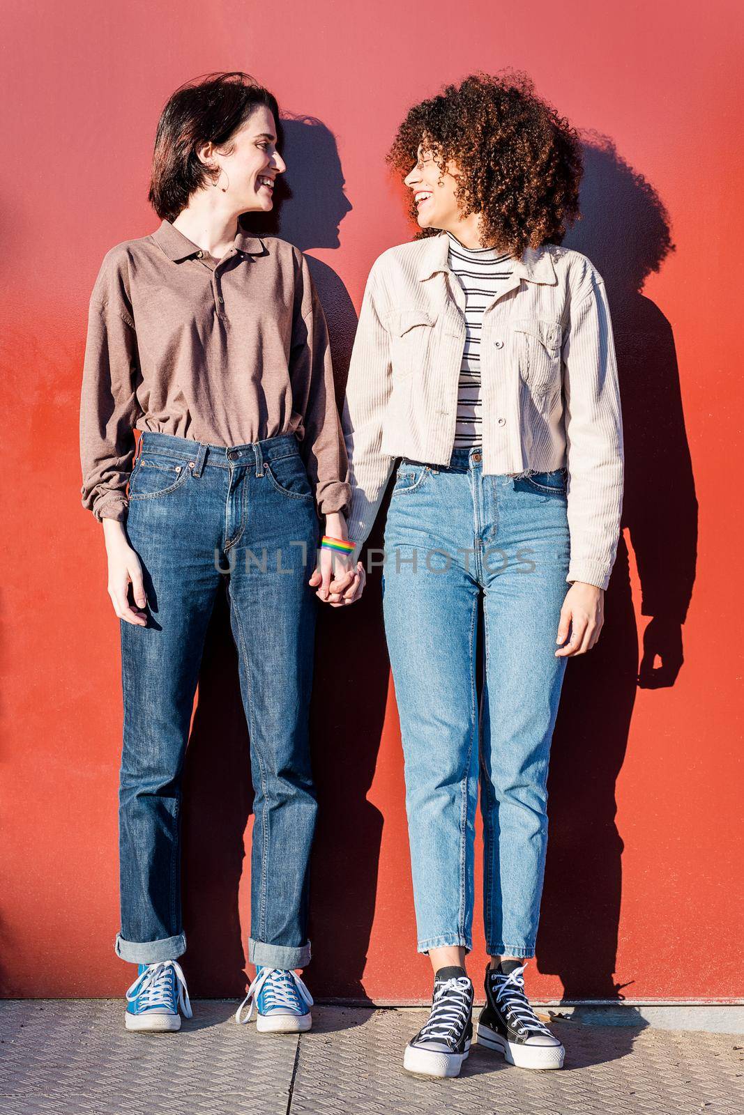 young interracial gay female couple looking at each other holding hands in front of a red wall, concept of sexual freedom and racial diversity, vertical photo