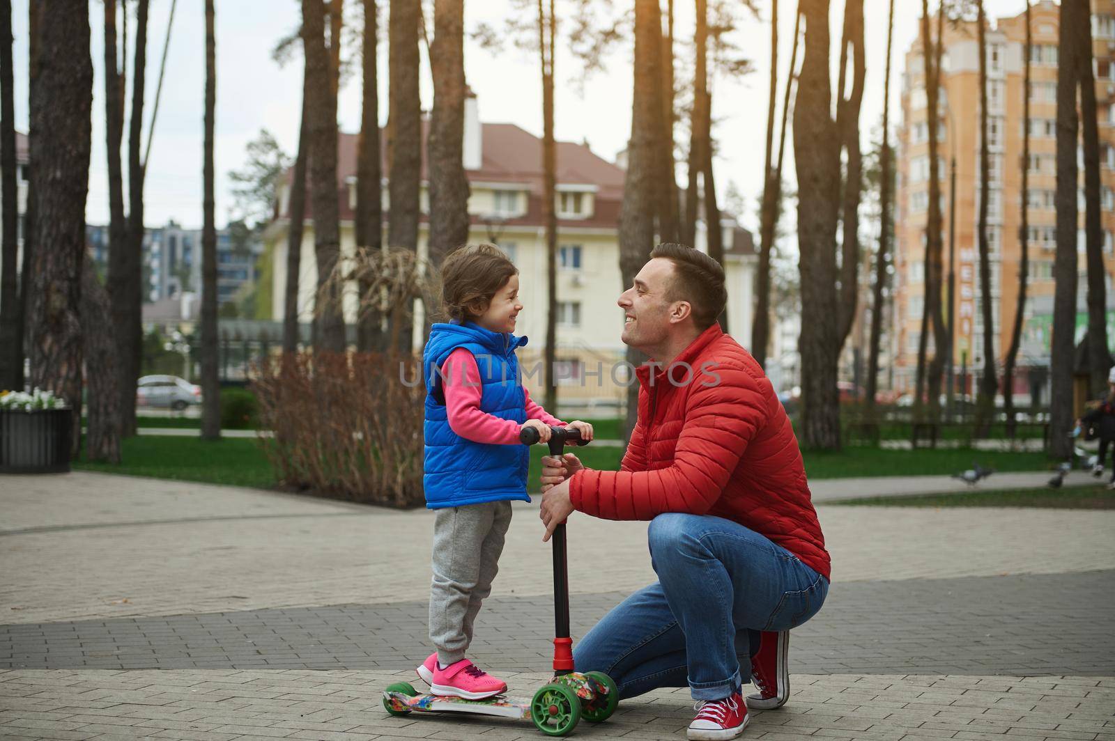 Side view of an adorable little girl on a kick scooter smiling cheerfully while looking at her father standing face to face with him in a city park on a beautiful sunny autumn day by artgf