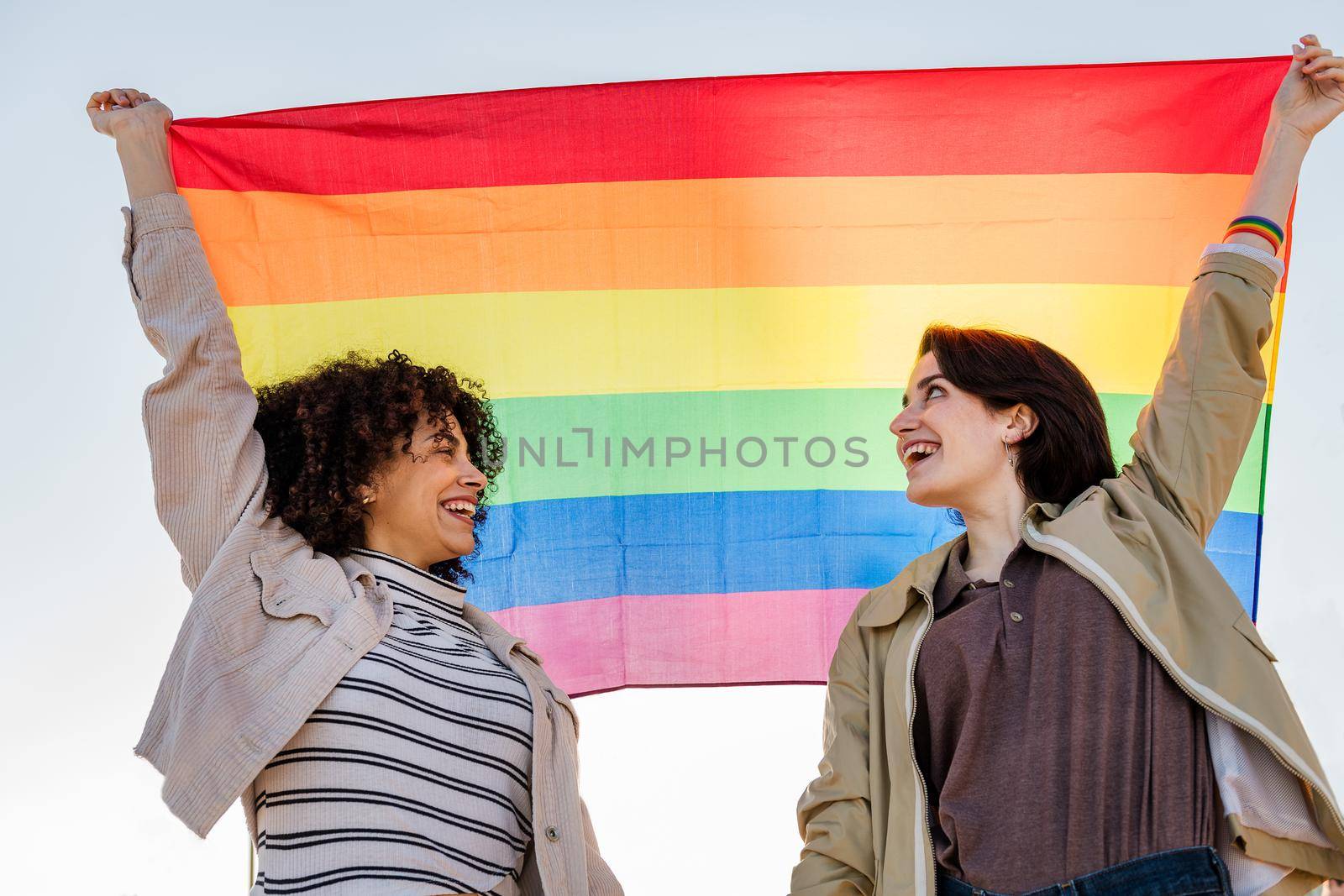 interracial homosexual couple of smiling women raising the rainbow flag, symbol of the struggle for gay rights, concept of sexual freedom and racial diversity