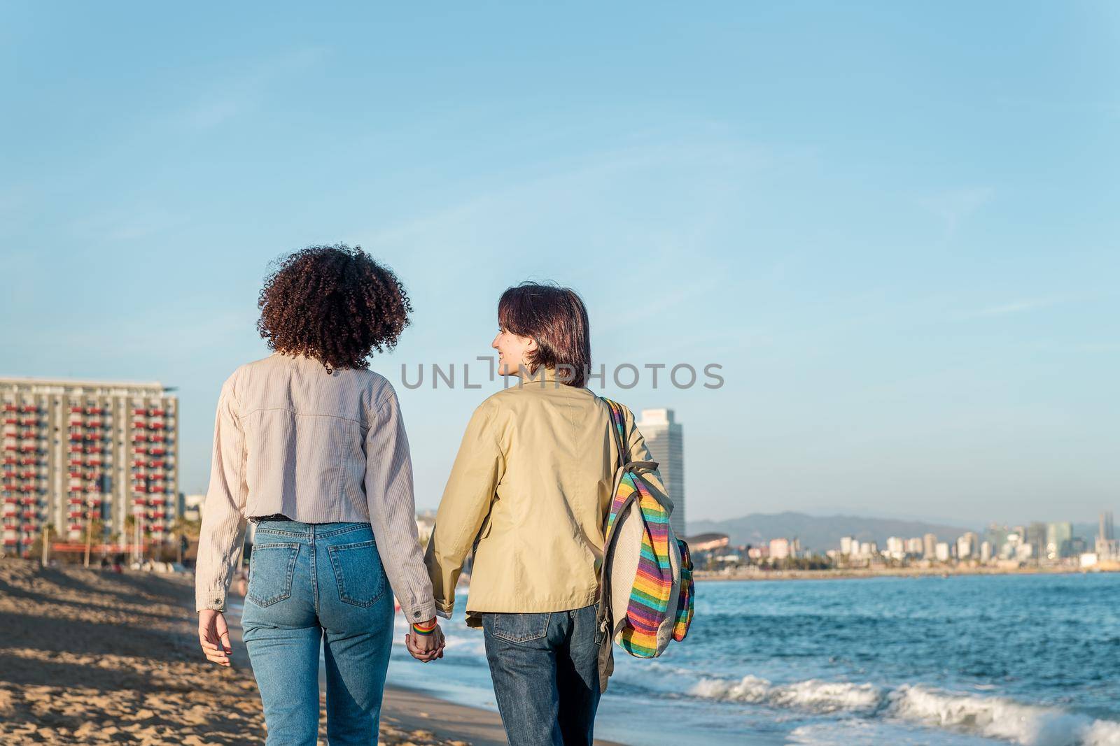 young couple of lesbian women walk hand in hand on the beach of Barceloneta in Barcelona, gay friendly city, with the colors of the rainbow flag, concept of sexual freedom and racial diversity