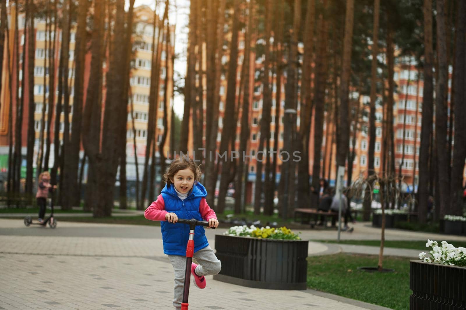 Adorable little Caucasian girl riding a kick scooter in the city park on a sunny day in early autumn or spring. Healthy and active lifestyle, leisure activity, happy childhood concept