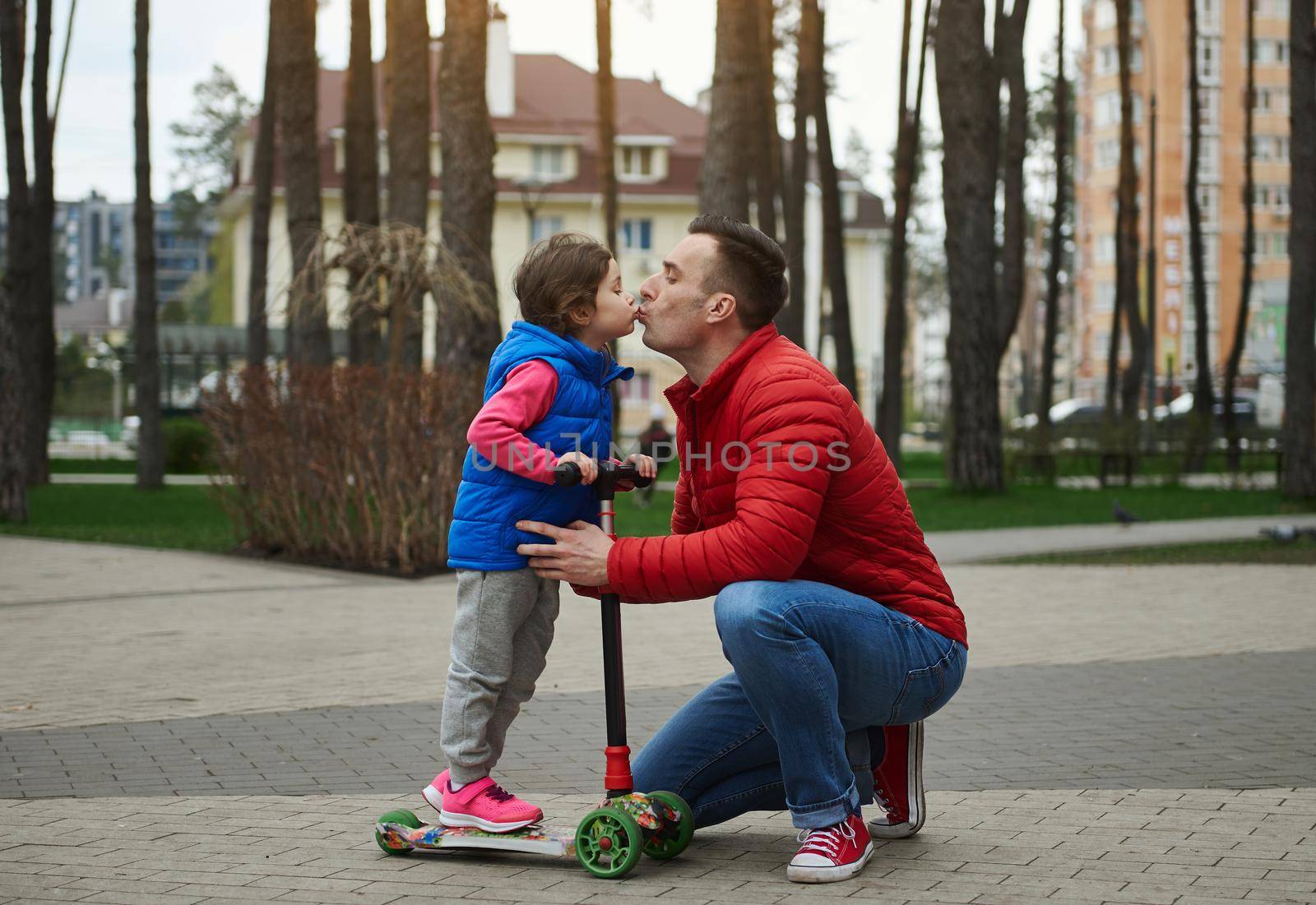 Loving caring dad and his beloved daughter on a push scooter in city park. Kisses, tenderness, love, care, affection togetherness, family relationships, childhood and fatherhood concept. Father's Day