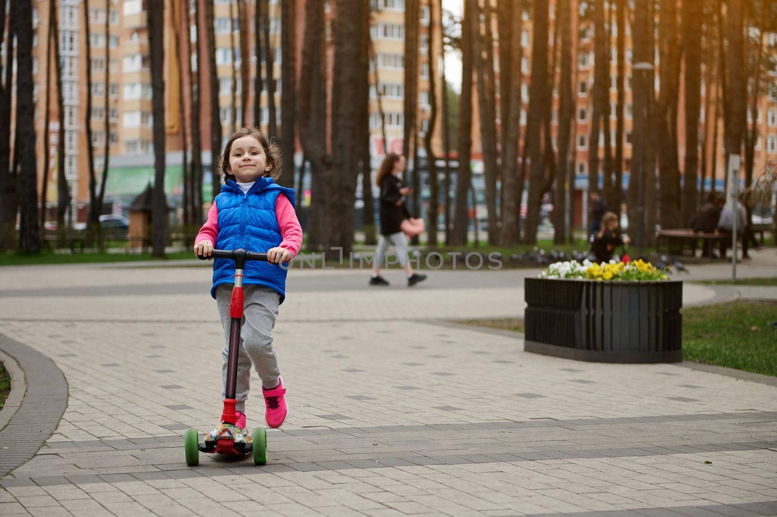 Full length portrait of a Caucasian cute little girl riding a kick scooter in the city park on a sunny day in early autumn or spring. Healthy and active lifestyle, leisure activity