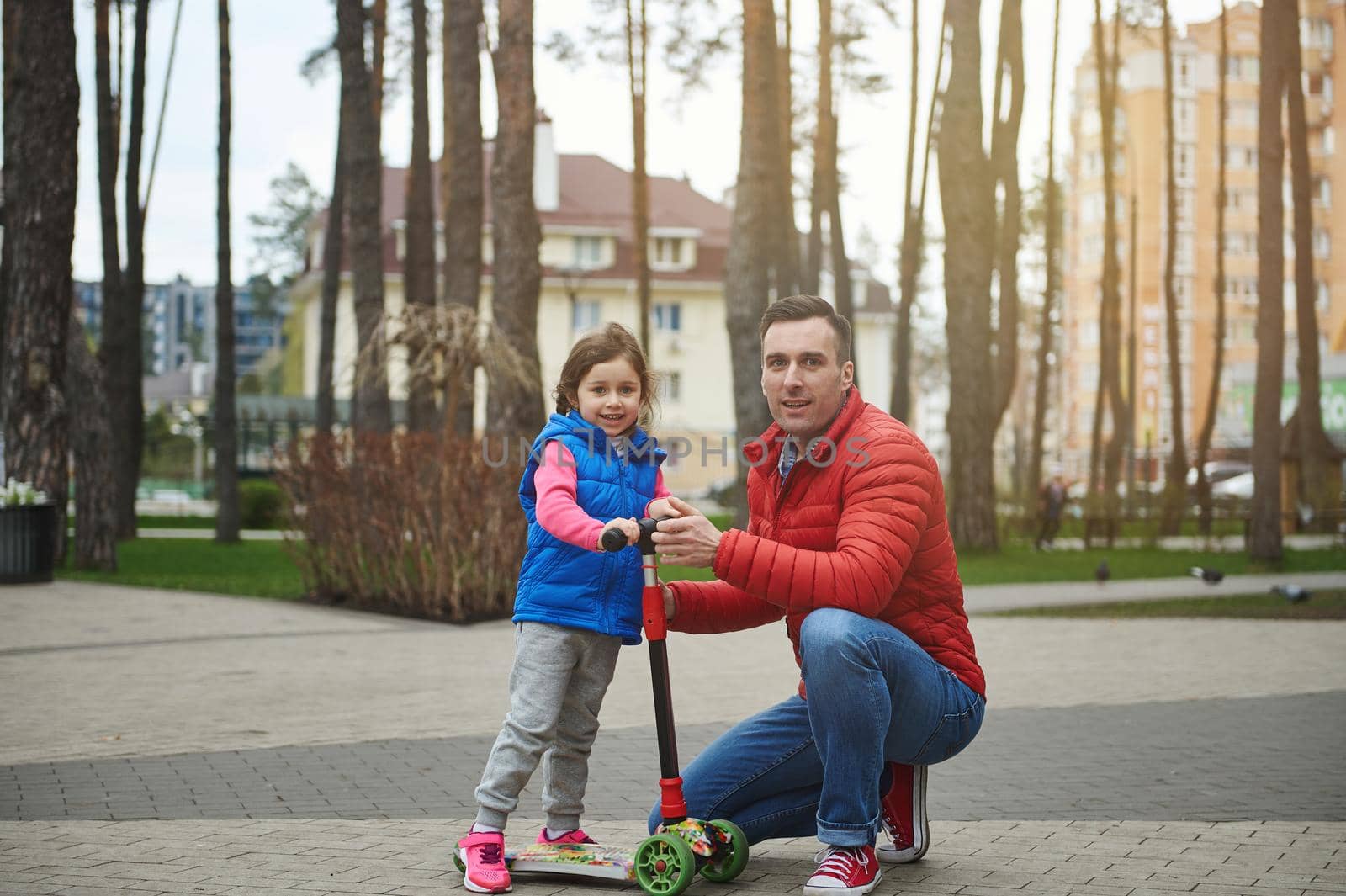 Handsome middle aged European man, happy caring father and his beloved daughter on a kick scooter, spending time together in an urban park by artgf