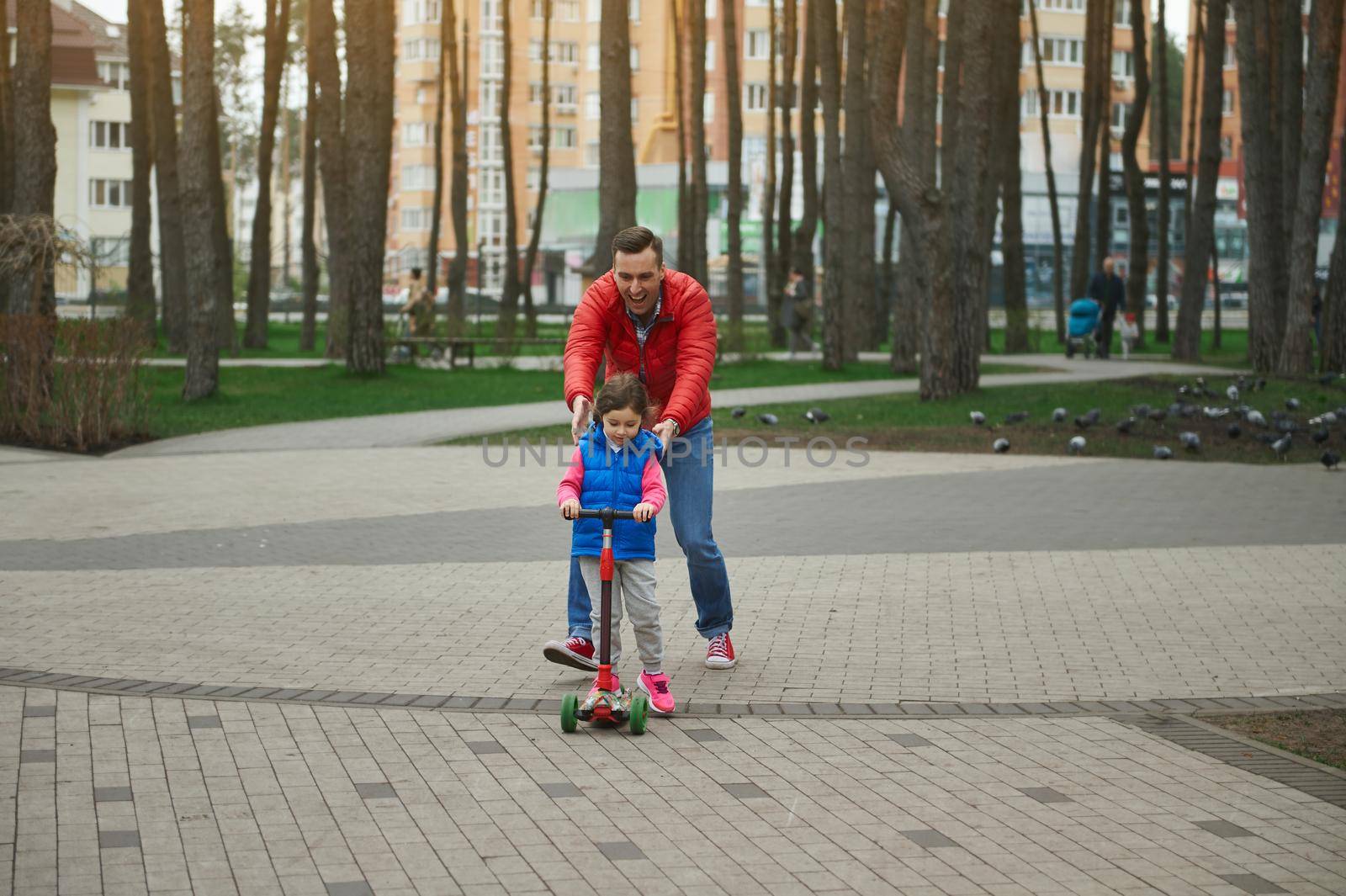 Cheerful dad running beside his cute daughter riding a push scooter, enjoying day off in a city park. Father's Day, happy childhood concept by artgf